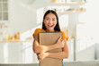 © Prostock-studio - In a warmly lit room, an excited asian woman joyfully hugs a carton box. She looks directly at the camera, radiant with happiness after receiving her parcel, feeling satisfied with her gift.