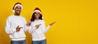 © Prostock-studio - Two cheerful individuals wearing white sweaters and Santa hats stand against a bright yellow background, pointing with smiles. They joyfully celebrate the Christmas season together.