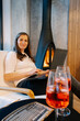 © Doralin - Woman working on laptop by fireplace with drinks