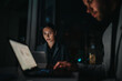 © qunica.com - A professional woman in a pinstripe suit studies data on a laptop in a dark, modern office, as a colleague types nearby. The scene conveys focus, teamwork, and late-night work stress.