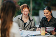 © qunica.com - Three people sit at a round table outside, smiling as they review papers and notes. A casual business meeting among friends and colleagues, with drinks and a notebook in a relaxed setting.