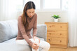 © KMPZZZ - Health care of knee pain concept, suffering asian young woman arthritis sitting joint ache, sore cramp or sprain tendon in leg on sofa at home. Osteoarthritis chronic disease of body problem, recovery