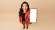 © Atmospheres - Smiling Asian woman holding a smartphone with a blank white screen for mockup, high angle view of a happy professional on a beige studio background.