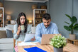 © Srdjan - Couple reviewing household budget at home woman with coffee and tablet, man focused on laptop; paperwork on table in cozy living room, finances, bills, savings, remote work.