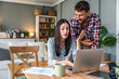 © Srdjan - Young couple working on the household budget. She explains numbers on the laptop while he checks his phone. Charts, coffee and documents on the table. Bills, savings, planning.
