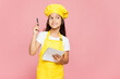 © ViDi Studio - Small kid child girl housewife chef cook baker mother's helper wear yellow apron toque cap write down in notebook recipe point finger up isolated on pastel plain pink background. Cooking food concept.