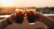 © kamral - Two hands clinking glasses of iced dark beverages against a warm, golden sunset sky during a celebratory moment.