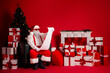 © deagreez - Santa sits in a red festive room reading a long list with gifts around a Christmas tree and warm holiday decor