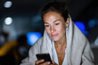 © Jovana - A woman wrapped in a soft blanket uses her smartphone at night in a relaxed setting, radiating calmness and emotional connection in a cozy atmosphere filled with soft lights.