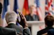 © Seventyfour - Black man raising hand during international conference, sitting among middle aged and senior Caucasian men and women, listening to speaker at podium with national flags in background