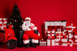 © deagreez - Santa sits amid gifts and a Christmas tree beside a red backdrop with a scooter and fireplace scene for a festive holiday stock image