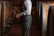 © 24K-Production - A man in a gray apron and shirt stands in a wine cellar, holding a glass of red wine near oak barrels.