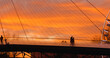 © RecCameraStock - Silhouetted individuals traversing transparent pedestrian bridge during golden hour sunset, capturing urban architectural elegance against dramatic orange sky