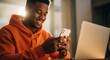 © Anna - Happy young Black man in an orange hoodie using a smartphone. Male student or freelancer working remotely from home with a laptop in a sunlit room