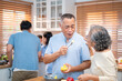 © Thitiporn - Asian elderly couple enjoying sweet kitchen moments while preparing food. The husband lovingly feeds his wife fruit, reflecting care, companionship, and joyful aging in a warm home.
