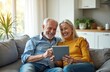 © Vadym - Elderly couple sits on couch laughing while looking at tablet. Husband and wife browse web on digital device together. Relaxed seniors enjoy leisure time indoors at home, sharing content online.