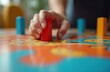 © Vadym - Person hand carefully moves bright red game piece on vibrant, colorful board. Strategic decision future business progress, financial planning, risk management, personal achievement. Fun leisure
