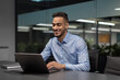 © Prostock-studio - A cheerful young middle eastern manager sits at a worktable in a modern office, focused on his notebook as he sends emails and conducts marketing research. The office has a relaxed atmosphere.