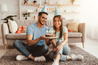 © Prostock-studio - A young couple sits on the carpet in their cozy living room, smiling and celebrating a birthday. They enjoy a delicious cake with candles while wearing party hats.