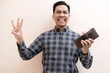 © Nurma - Southeast Asian man wearing flannel shirt smiling happy holding leather wallet with giving number 12345 by hand gesture, on white isolated background.