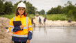 © Happy Photo - A woman in a hard hat and safety jacket stands by a waterway, holding a clipboard. In the background, workers are seen engaging in construction activities. It is daylight and the site looks busy