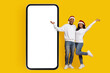 © Prostock-studio - A cheerful couple wearing Santa hats stands next to a large blank smartphone screen. They pose with smiles and raised arms, celebrating the festive season in a vibrant yellow setting.