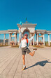 © EdNurg - Happy tourist holding small Kazakhstan flag and jumping in front of the arch in First President Park, Almaty