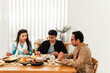 Three people share a home-cooked meal at a bright dining table, with rice, fruit and curries, chatting in a cozy, sunlit room; two wear traditional attire