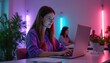 © Vadym - Young woman works on laptop in modern office with neon lights. Female sits at desk typing on computer keyboard. Plants are on table and around office space. Another woman is working in background.