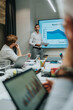© qunica.com - A business presenter stands beside a large display showing results while colleagues work on laptops in a bright conference room. A collaborative session focused on data, strategy, and planning.