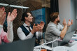 © qunica.com - A group of people sits around a conference table, clapping and smiling in a bright office. The scene conveys teamwork, motivation, and positive momentum in a business meeting or project kickoff.