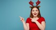 © Cleverson - Young woman wearing reindeer antlers and a red shirt, pointing to the side with a surprised expression on a blue background