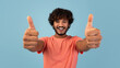 © Prostock-studio - A handsome young eastern guy in a pink t-shirt poses alone against a blue background. He smiles warmly while giving a thumbs up, suggesting he recommends something positive.