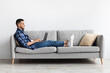 © Prostock-studio - Young Arab man sits comfortably on a sofa in his living room, smiling while browsing the internet on his laptop. Casual setting with a relaxed atmosphere for personal enjoyment.