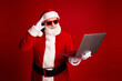 © deagreez - Santa in red costume with sunglasses holding a laptop against a bold red background perfect for holiday marketing and Christmas themed stock photos