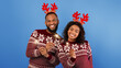 © Prostock-studio - Celebration time. Joyful black couple in deer horns and identical sweaters celebrating Christmas or New Year with sparklers over blue studio background