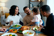 © AnnaStills - Man holding toddler girl while sitting at dining table with Black woman and another man, all smiling and engaging during family meal with roast chicken and salad
