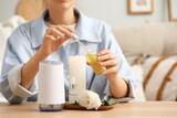 Woman with eustoma flower and candle adding essential oil to air humidifier on table in room, closeup