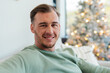 © Wavebreak Media - Mid-adult man relaxing on light sofa at home, gazing toward window showing decorated Christmas tree