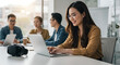 © Arrhman - Smiling woman working on laptop with colleagues in modern office setting.