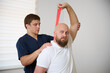 © Василь Івасюк - Physiotherapist guiding male patient with resistance band exercise for shoulder rehabilitation in clinic. Physical therapy session focused on injury recovery, strengthening, mobility improvement