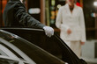 © AnnaStills - Caucasian man in suit wearing glove opening car door for Caucasian woman in business attire approaching vehicle, both appearing as middle aged professionals in urban setting