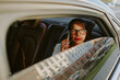© AnnaStills - Middle aged Caucasian woman sitting in back seat of car talking on smartphone, wearing glasses, looking out window with thoughtful expression, city street visible outside