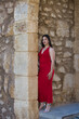 © @skuder_photographer - A beautiful teenage girl with long dark hair, dressed in red and wearing high heels, leaning under a Roman archway on a street in a typical Andalusian town. In the background is a wall.