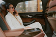 © AnnaStills - Middle aged Caucasian woman sitting in back seat of luxury car talking on smartphone, wearing glasses, short hair visible, city buildings seen through window