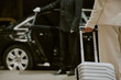 © AnnaStills - Caucasian middle aged man holding suitcase standing near luxury car while chauffeur in uniform opening door, business travel concept, partial body view, no faces visible
