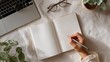 © Natalia S. - Top view of female Asian hand holding pen over open blank notebook near laptop and plants. Concept for planning, journaling, education, or creative writing workspace