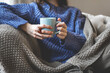 © Anna Zhukkova - Close up female hands holding blue cup of tea or coffee. Woman in knitted sweater and cozy plaid relaxing at home on sofa, warm and peaceful indoor atmosphere