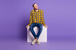 © deagreez - Young handsome man in plaid shirt sits on white block against purple background smiling up casual fashion lifestyle and positive vibe