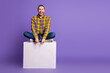 © deagreez - Happy young man in plaid yellow shirt sits on a white block against a purple background smiling and pointing down